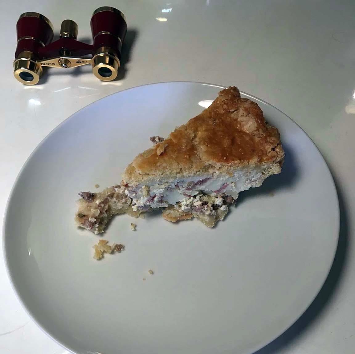A slice of Easter pie, golden on top and white on the sides, with crumbled sausage coming out at the bottom, sits on a white plate on a white table. Next to it, for some reason I can't remember, is a pair of opera glasses with a dark red body and gold-toned fittings.