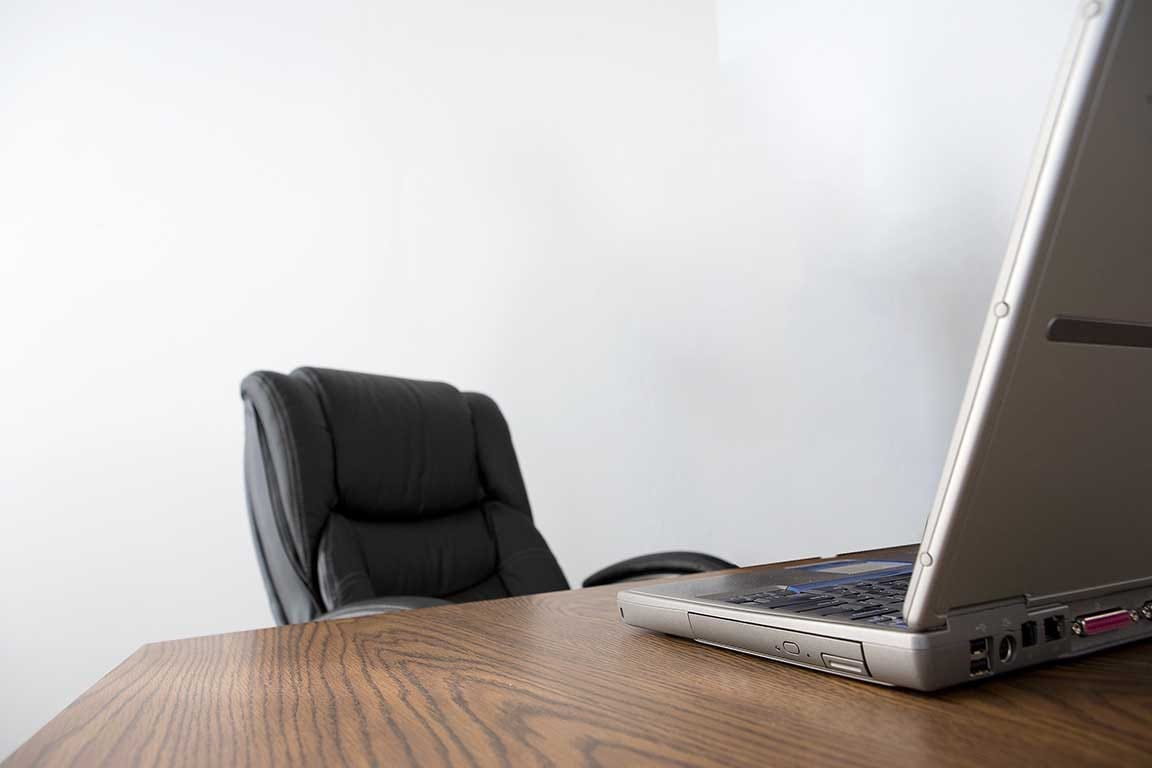 An empty office chair in a featureless room at a table in front of a laptop computer