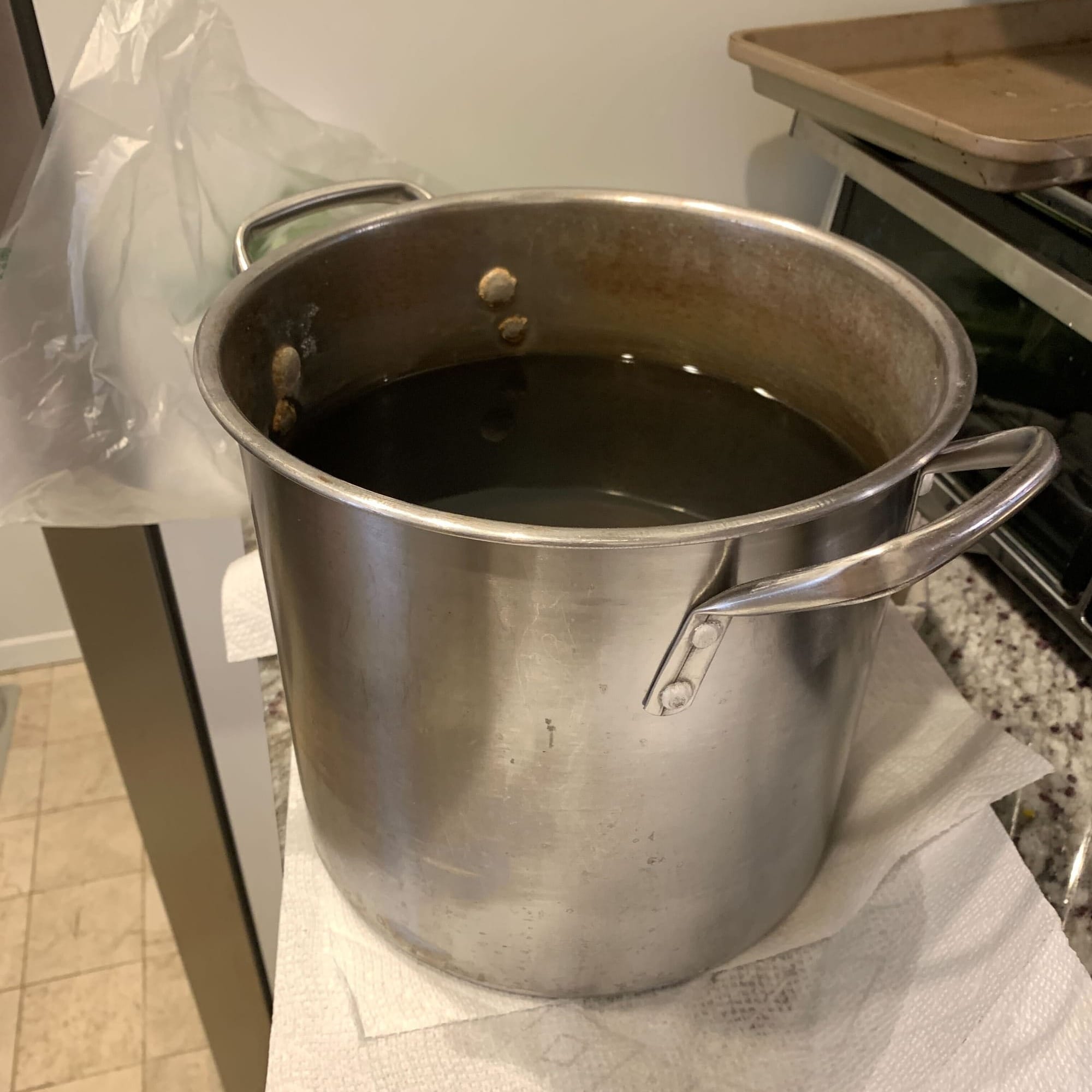 A battered steel pot with water just below its internal rivets sits on a damp paper towel on a granite kitchen counter. A toaster is visible to the right and a tile floor to the left, and there's a clear plastic bag sticking up behind the pot. 