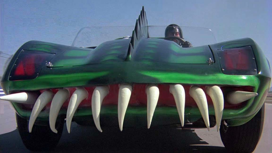 A weird green car painted up like a giant reptile and it has scraggly giant pointy teeth for a grille