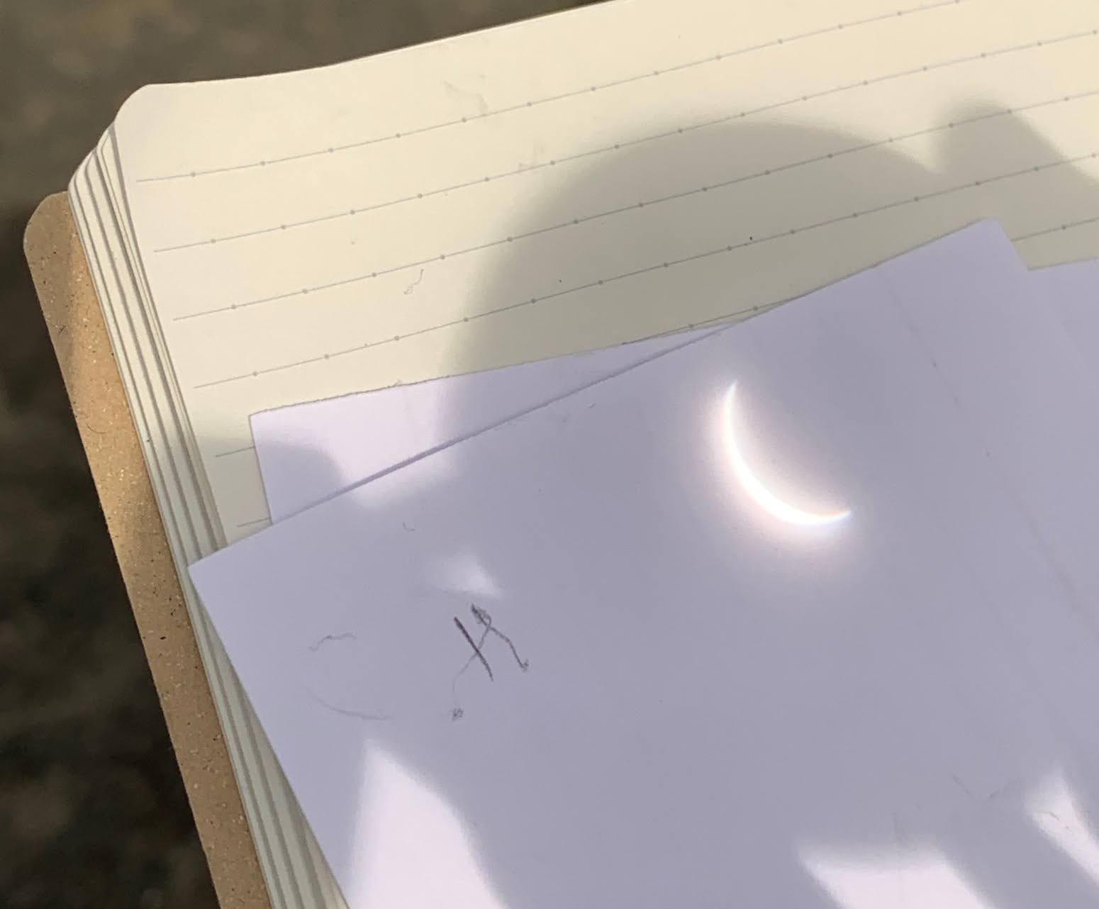 Alt: The crescent sun is projected through a monocular onto a white card resting on a lined notebook page. The shadow of the monocular surrounds the image of the sun.