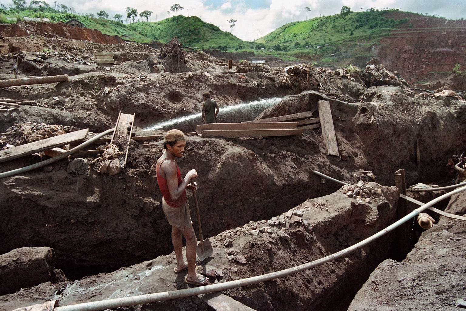 Emerald green hills behind a completely ripped up landscape a man with a shovel in foreground, and a figure in middle ground watching water spraying onto a trench.
