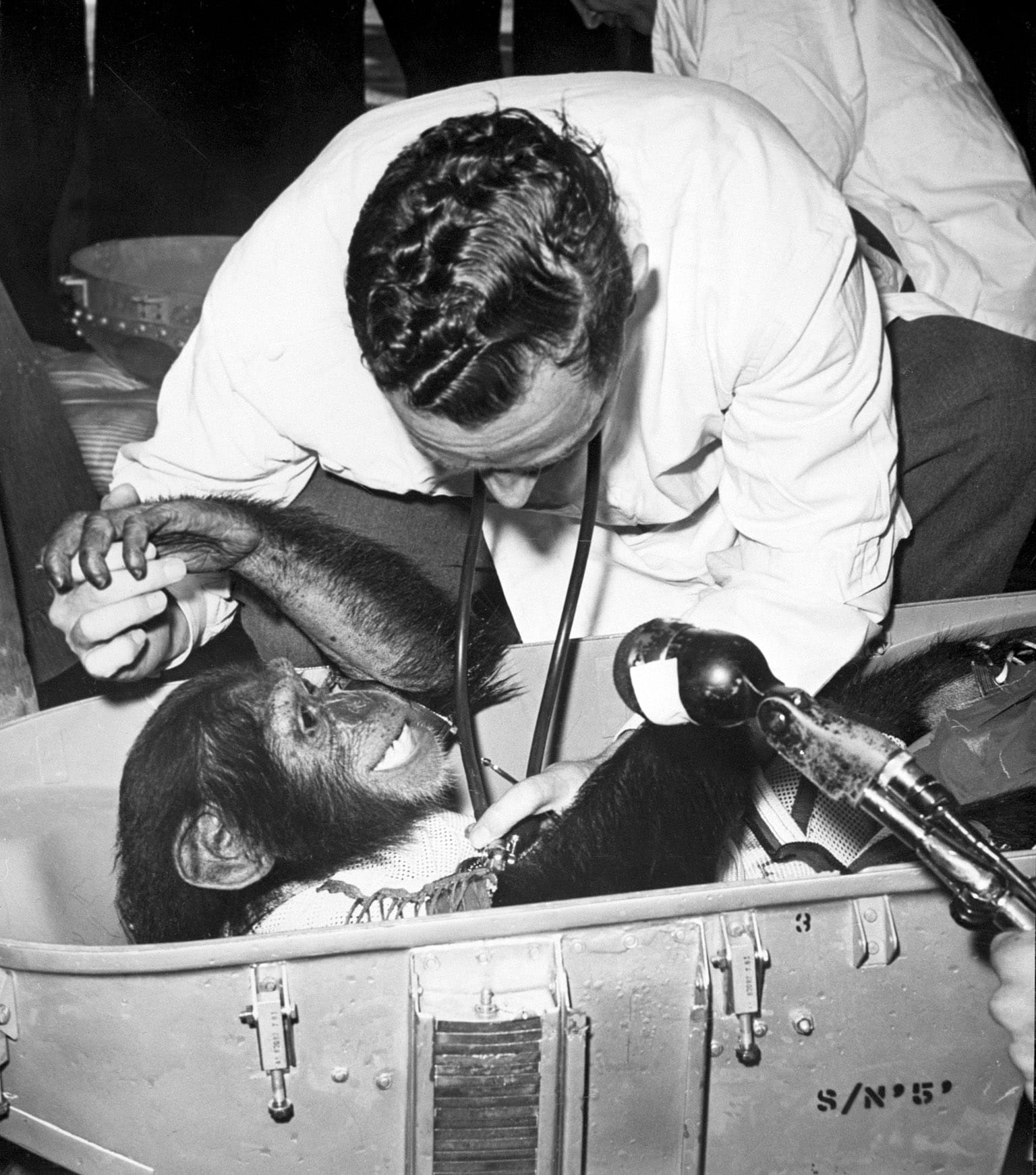 Ham, in his space container, looks sort of happy to be having his heart listened to by the guy with the stethoscope but they just shot his monkey ass into space, so he's likely in shock.