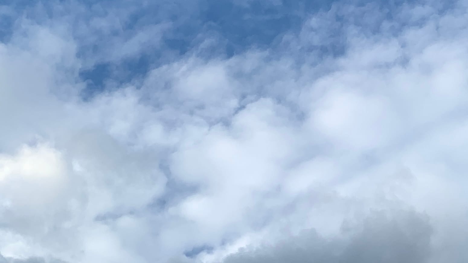 A sky mostly covered in cottony white clouds, with blue showing through them at the top of the frame and along a few other rifts, including a strong angled line from roughly center bottom to roughly center right, making a triangle in which darker gray clouds push up into the frame from the bottom. 