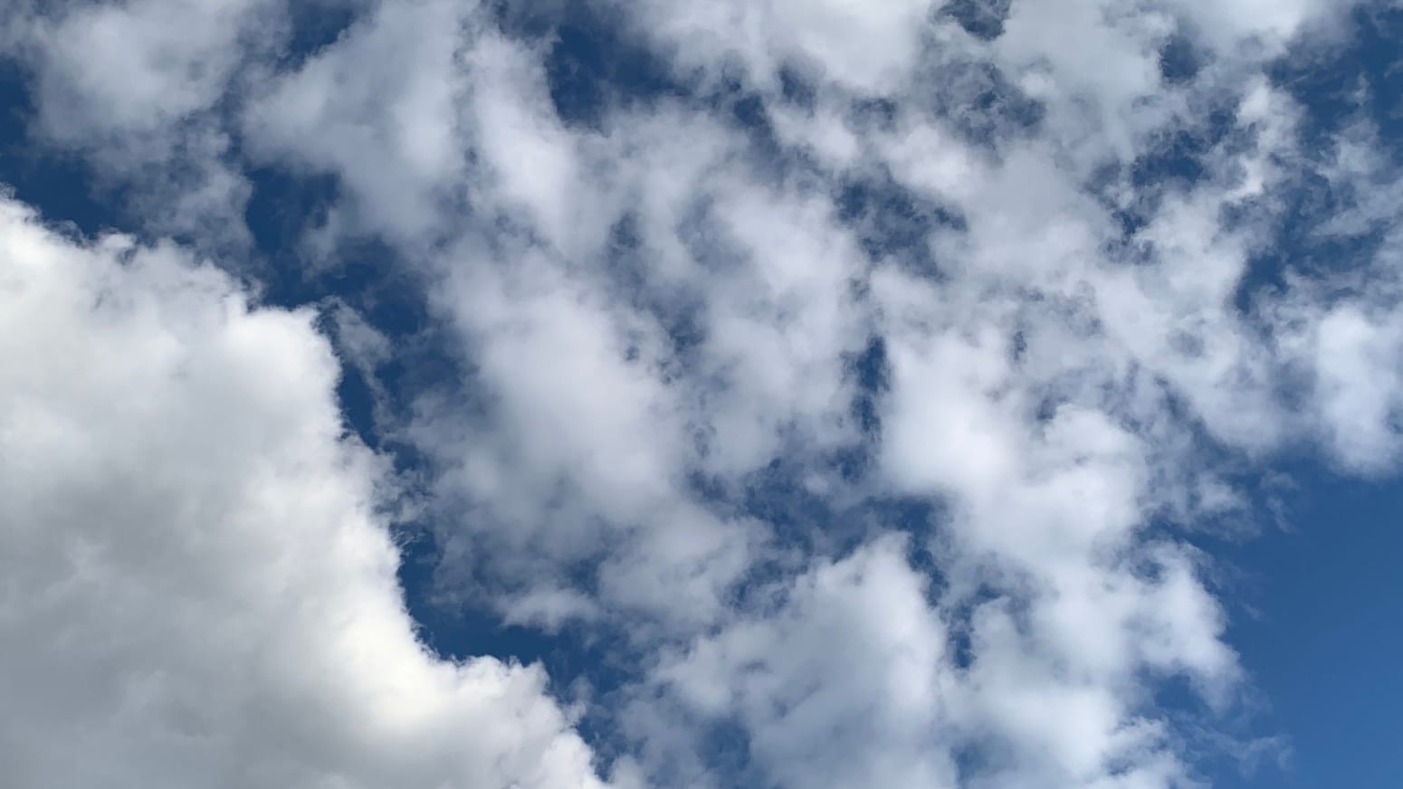 A sky of deep blue showing in gaps through broken white clouds, with a thicker, brighter white cloud in the lower left corner and an open patch of pure blue in the lower right corner. 