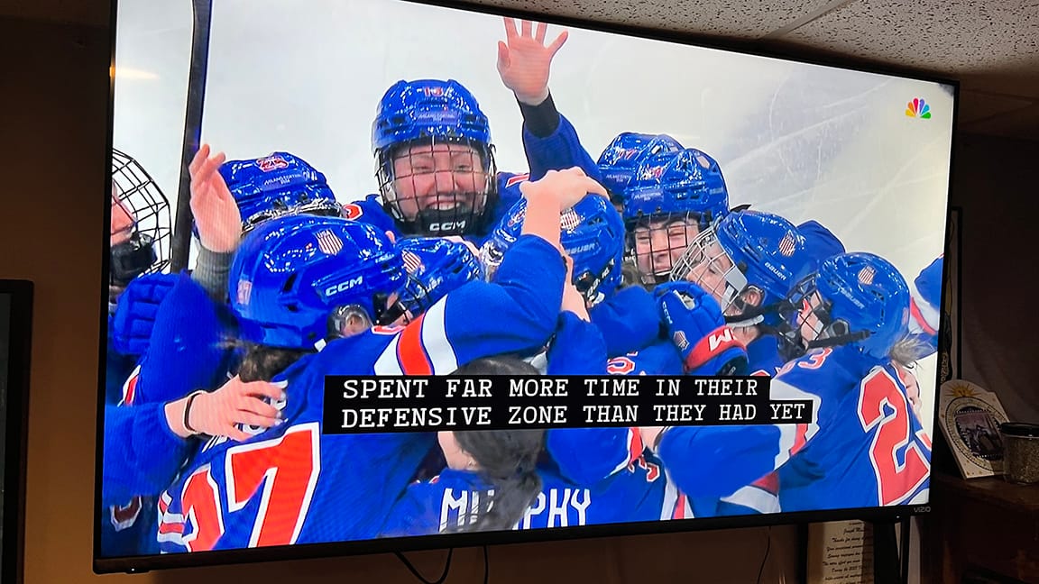 Photograph of a television on which Team USA in blue hockey uniforms forms a giant grinning hug-unit on the ice under a closed-caption display reading "SPENT FAR MORE TIME IN THEIR DEFENSIVE ZONE THAN THEY HAD YET"
