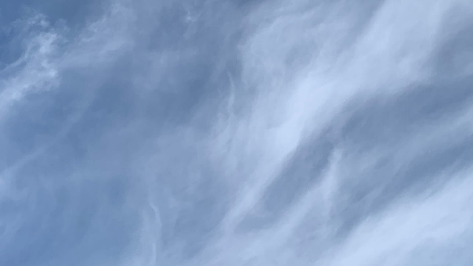 A patch of sky with complicated brushy clouds cutting this way and that at different angles, blurring over a blue background. There are some less brushy and more clumpy, but still translucent, bits of cloud in the top left corner.  