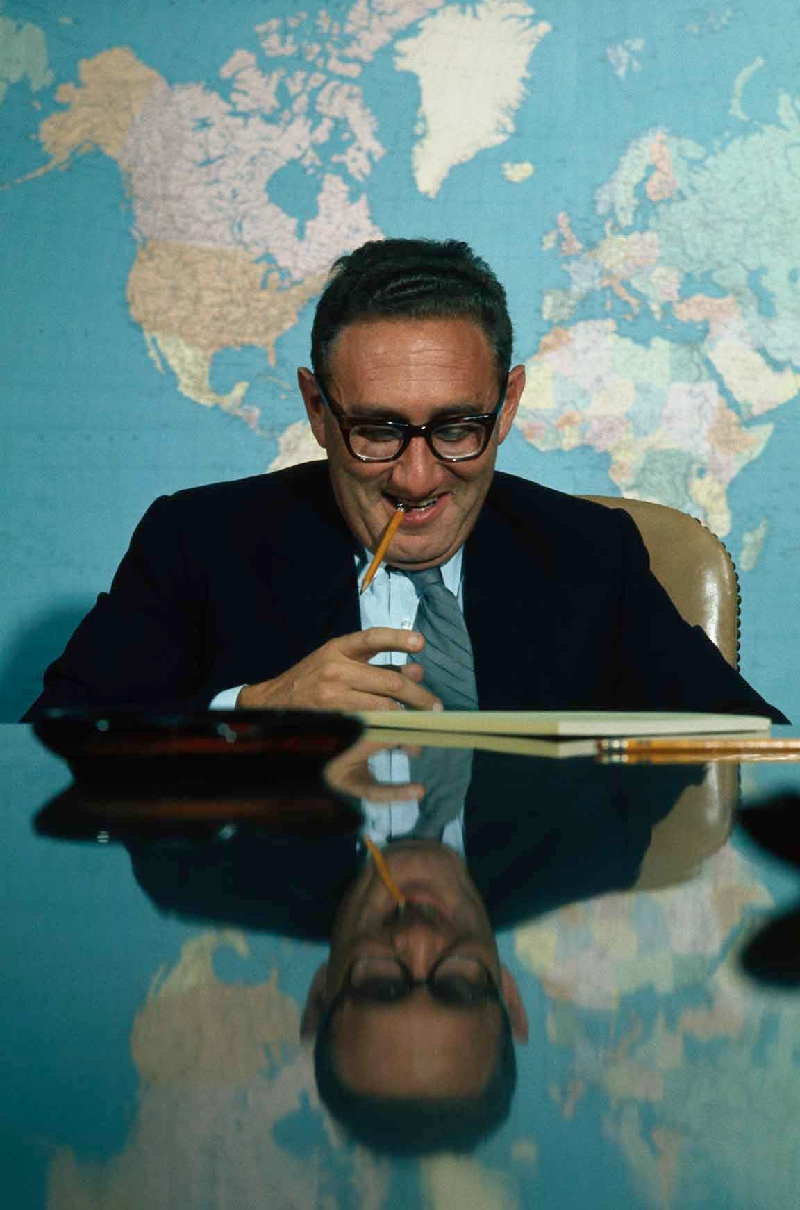Kissinger sitting at table with map of world behind him. Looking down, smiling, pencil dangling from his mouth.