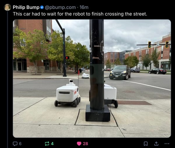 Three foodbots at an intersection and a car is waiting for one to cross the street