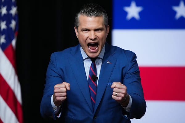 Man in blue suit near US flag with US flag backdrop making fists yelling mouth very open