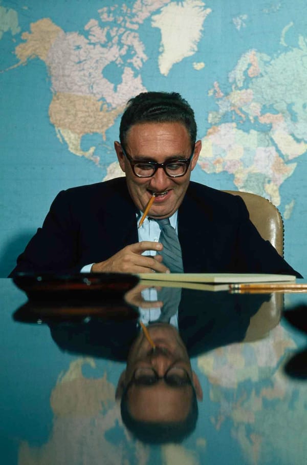 Kissinger sitting at table with map of world behind him. Looking down, smiling, pencil dangling from his mouth.
